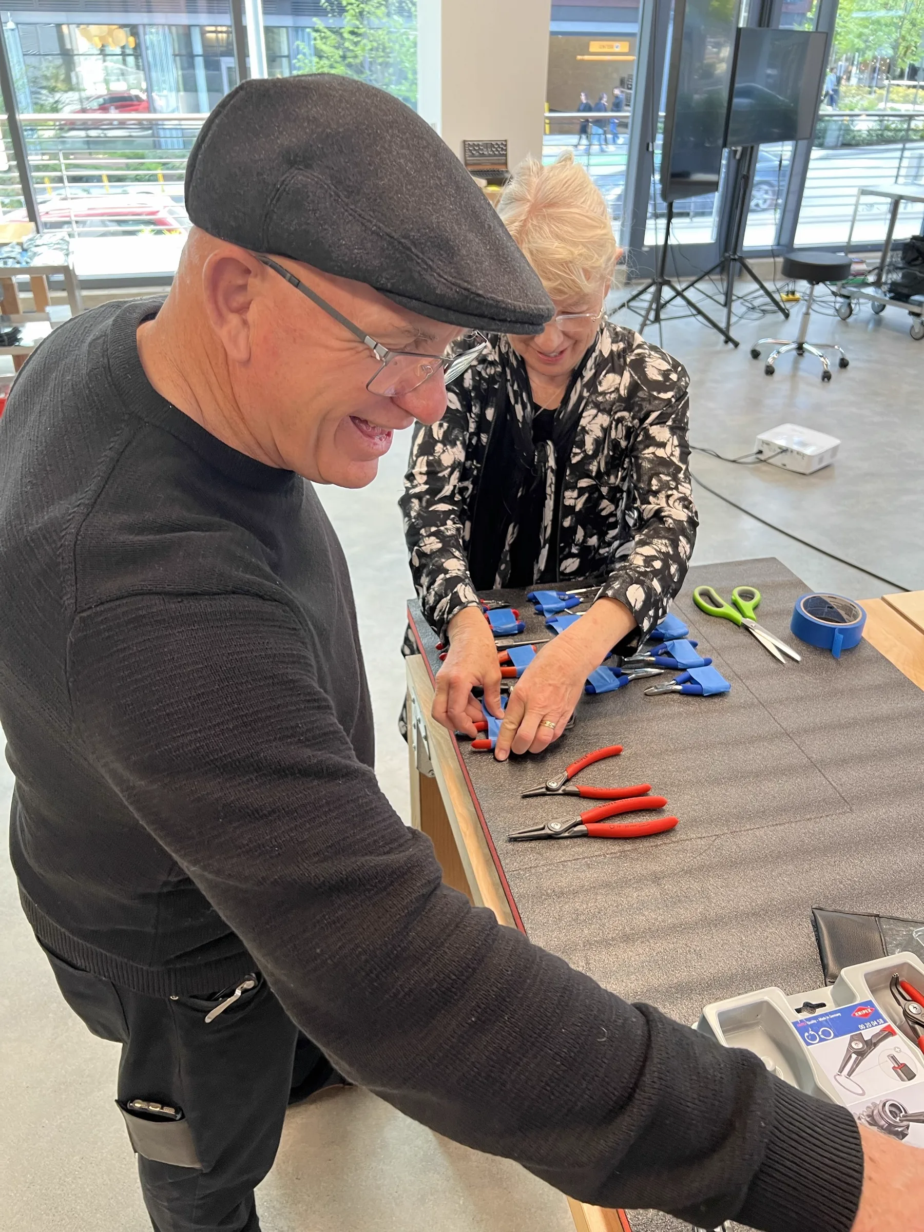 Joshua Borsman's parents at the studio workbench organizing pliers into a foam tool tray, South Lake Union visible through the glass