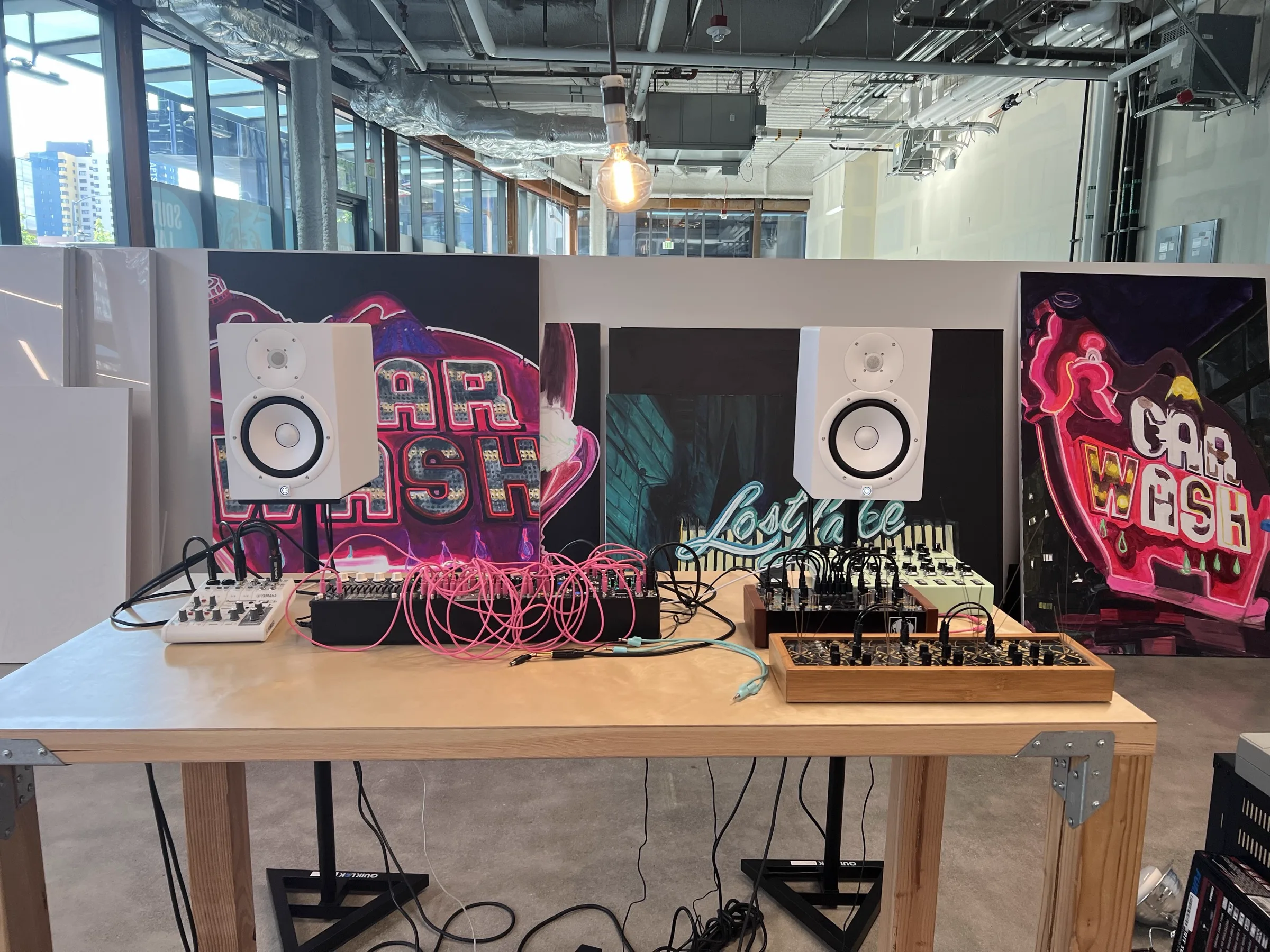 Studio workbench with modular synthesizer, monitors, Car Wash and Lost Lake paintings on the wall, Edison bulb overhead, South Lake Union skyline through the glass