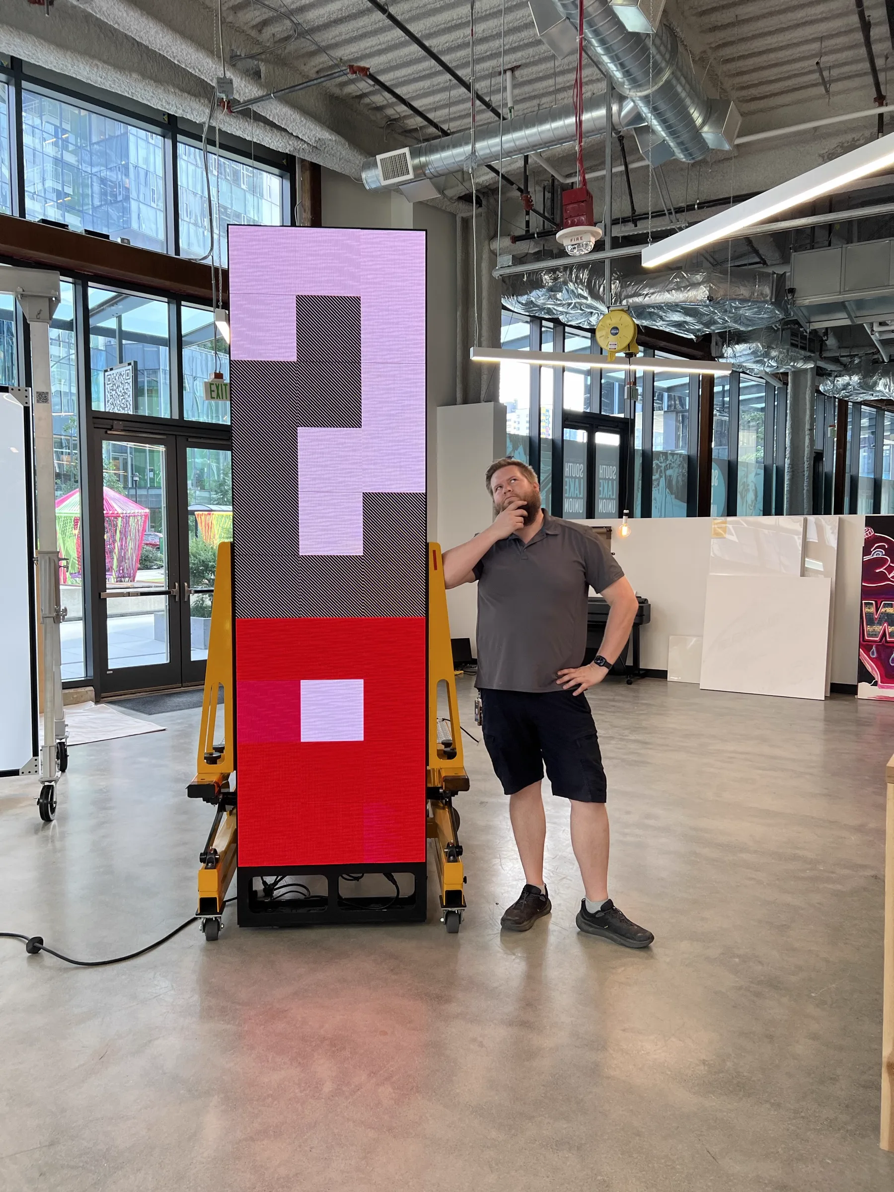 Joshua Borsman in the open studio working with a large pink-and-red LED pixel-art panel, South Lake Union streetscape visible through the glass
